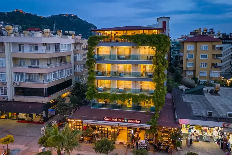 Gallion Hotel exterior at dusk in Alanya, with balconies above its ground-floor pizza and snack bar.