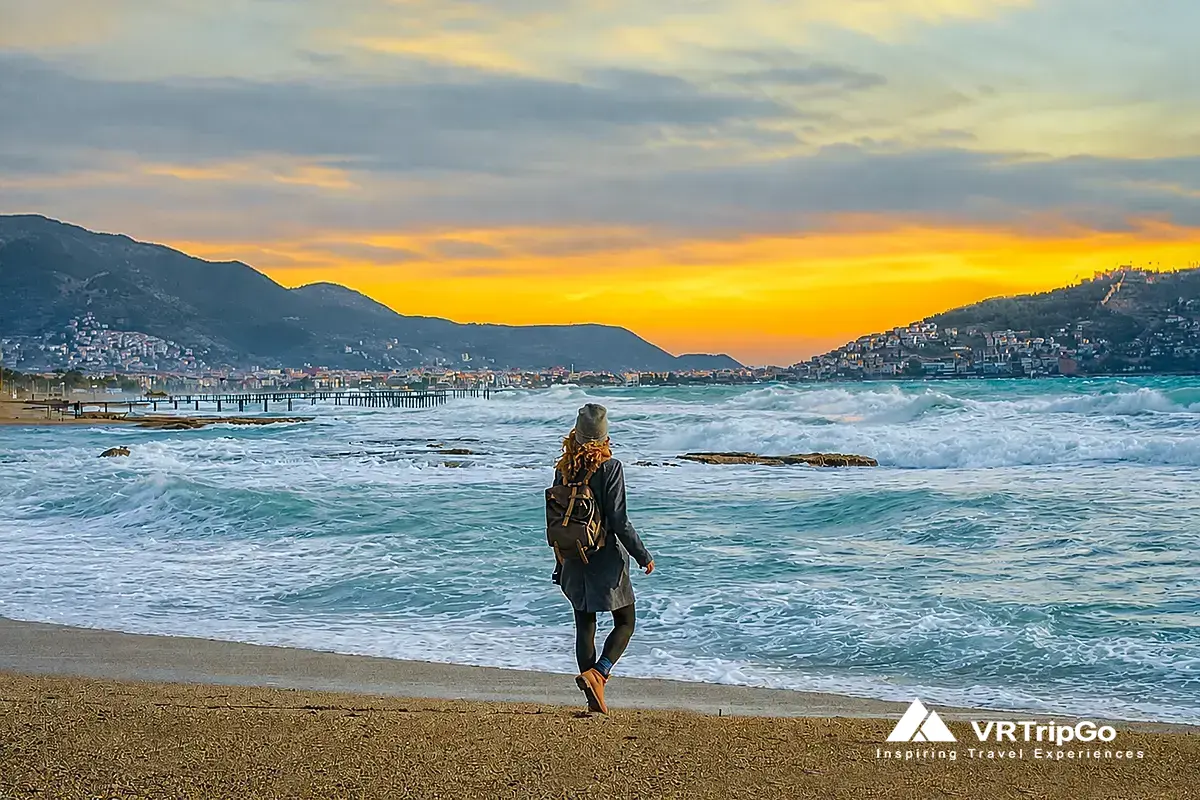 Woman walking along Kemer beach at sunset with waves, mountains and coastline, scenic view of Kemer Turkey travel destination