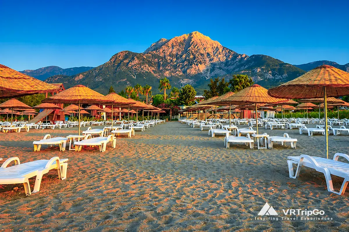 Sunny beach in Kemer during spring with clear sky, warm light, empty sunbeds and mountain backdrop, showing mild and pleasant spring weather in Kemer