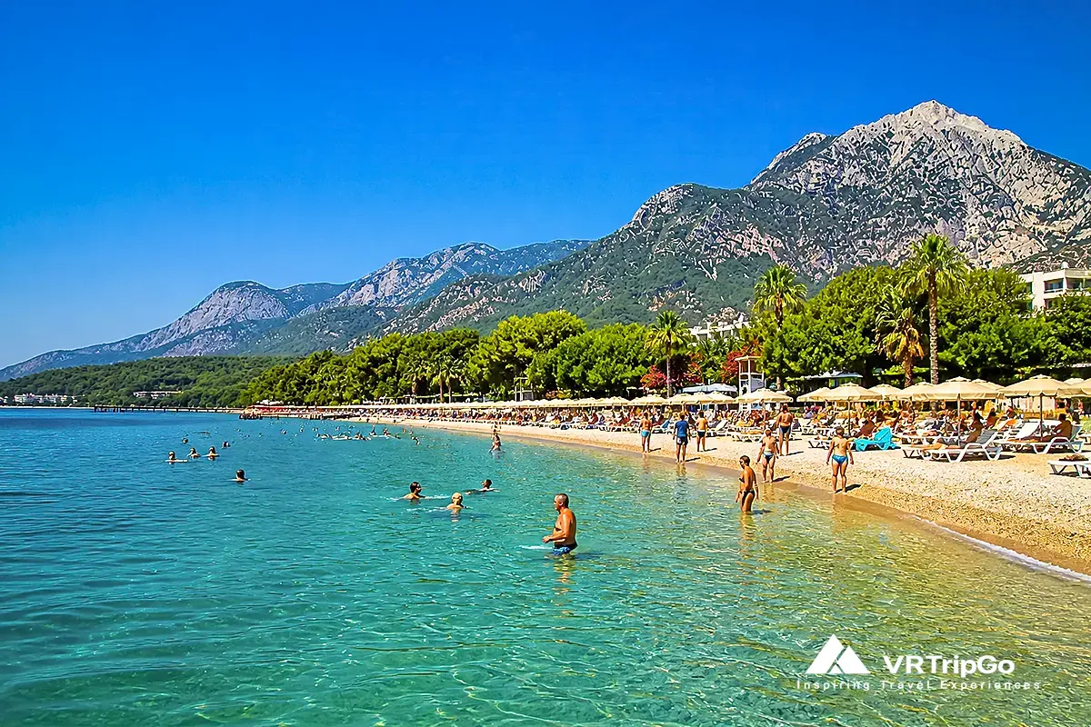People swimming in the clear sea in Kemer during summer with sunny weather, turquoise water, beach umbrellas and mountains in the background