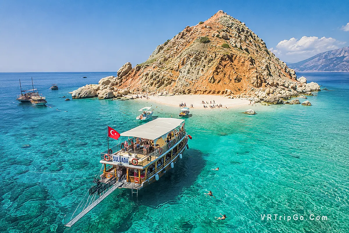Suluada Island Boat Tour featuring crystal-clear turquoise waters, excursion boats near the famous white beach of Suluada, and breathtaking Mediterranean island scenery on the Antalya coast.