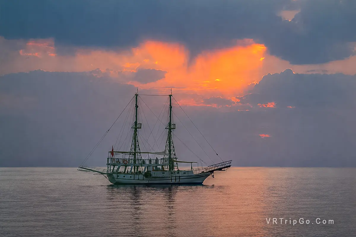 Bodrum Boat Tour featuring a traditional wooden yacht cruising across calm waters at sunset with tourists enjoying the scenic Aegean Sea views.