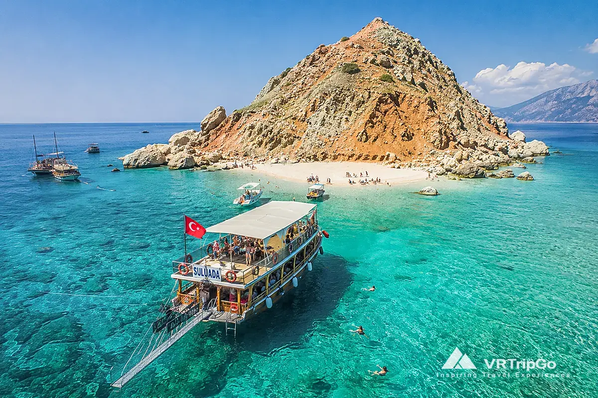 Suluada Island Boat Tour featuring crystal-clear turquoise waters, excursion boats near the famous white beach of Suluada, and breathtaking Mediterranean island scenery on the Antalya coast.