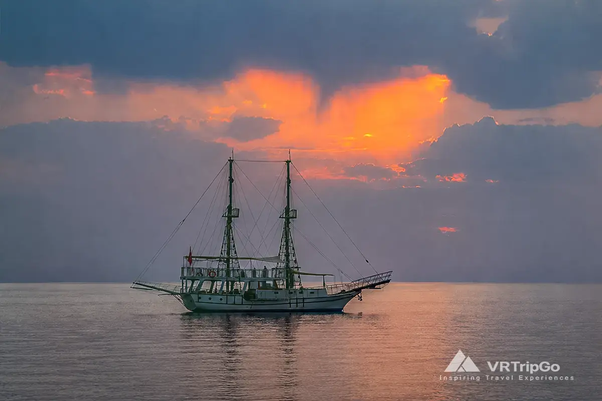 Bodrum Boat Tour featuring a traditional wooden yacht cruising across calm waters at sunset with tourists enjoying the scenic Aegean Sea views.