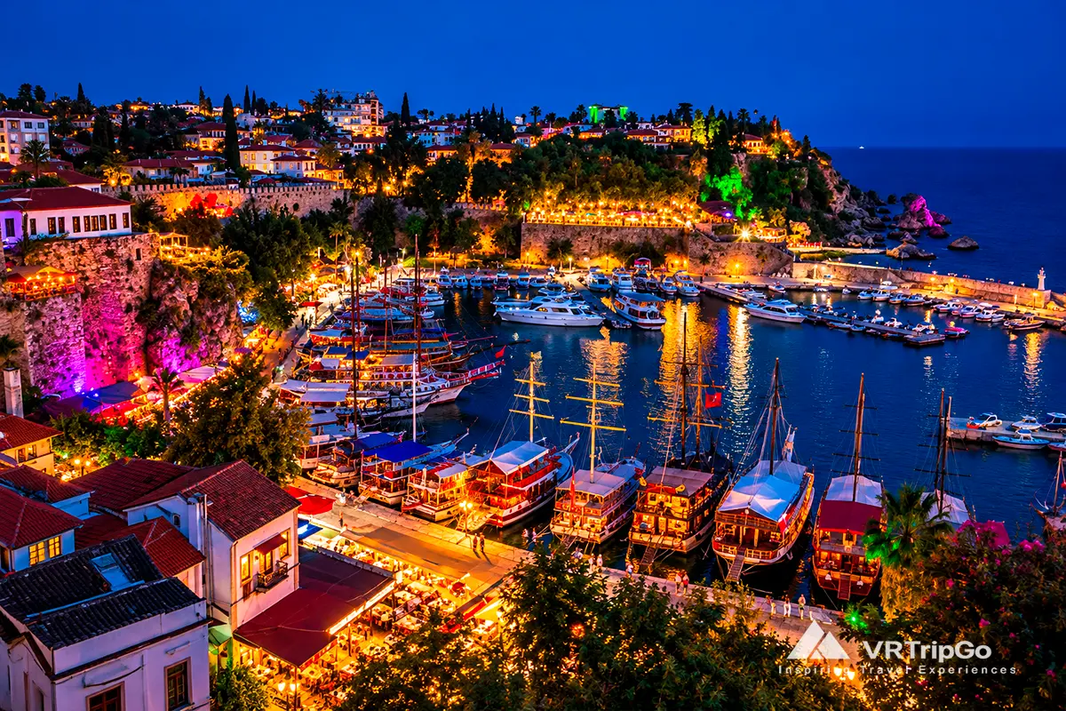 Panoramic evening view of Antalya Old Harbor in Kaleici, with illuminated historic buildings, yachts, tour boats, and colorful waterfront lights.